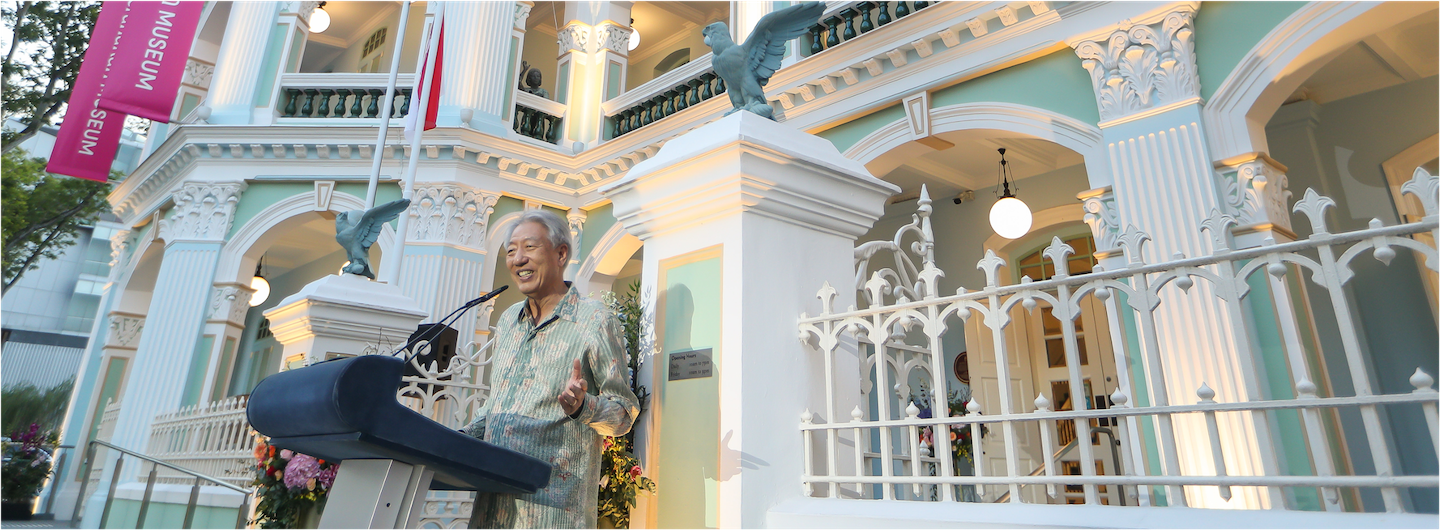 Man speaking at podium outside Peranakan Museum, with eagle statues and white fence.
